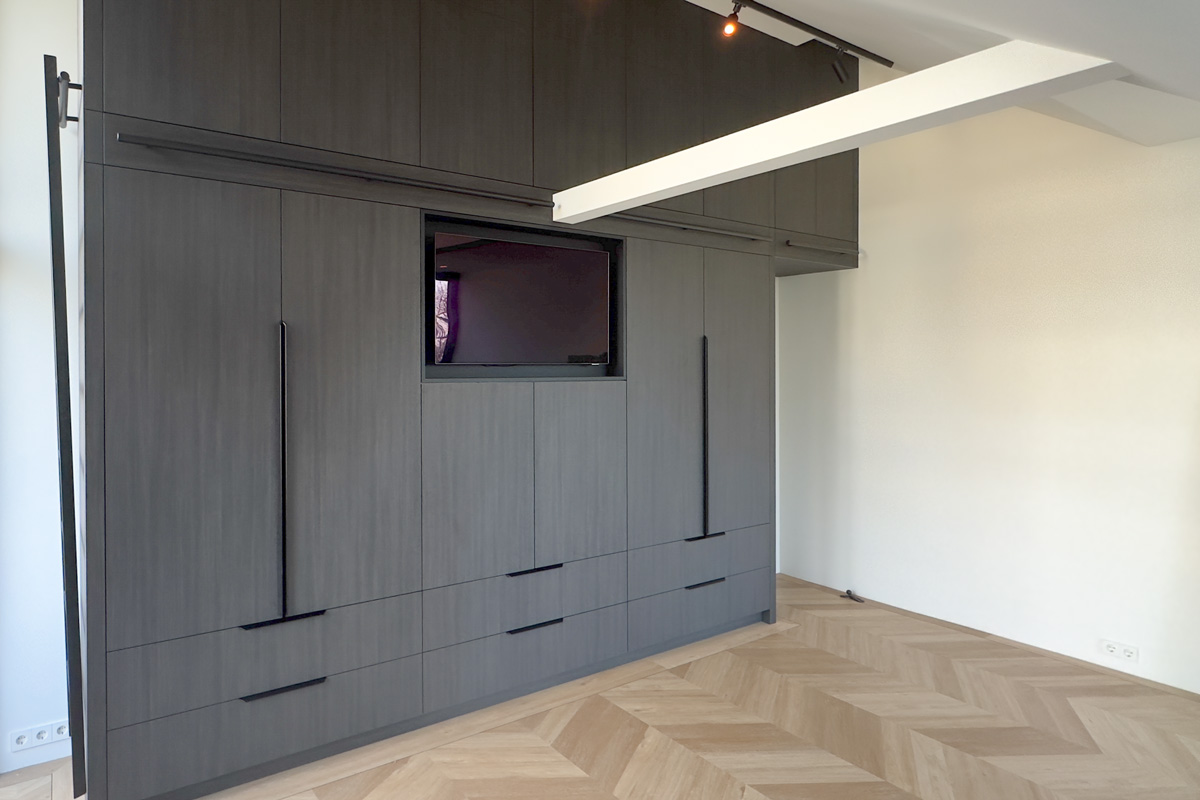 Donkere maatkast in een slaapkamer met strakke handgrepen, ingebouwde lades en een geïntegreerde nis voor een televisie, geplaatst op een visgraatvloer.