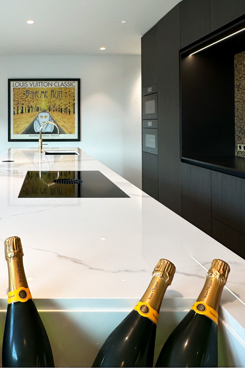 Detail van een grote luxe keuken met een marmeren werkblad, geïntegreerde champagnekoeler en een modern zwart wandmeubel met gouden accenten. Op de achtergrond een opvallend kunstwerk voor een unieke uitstraling.