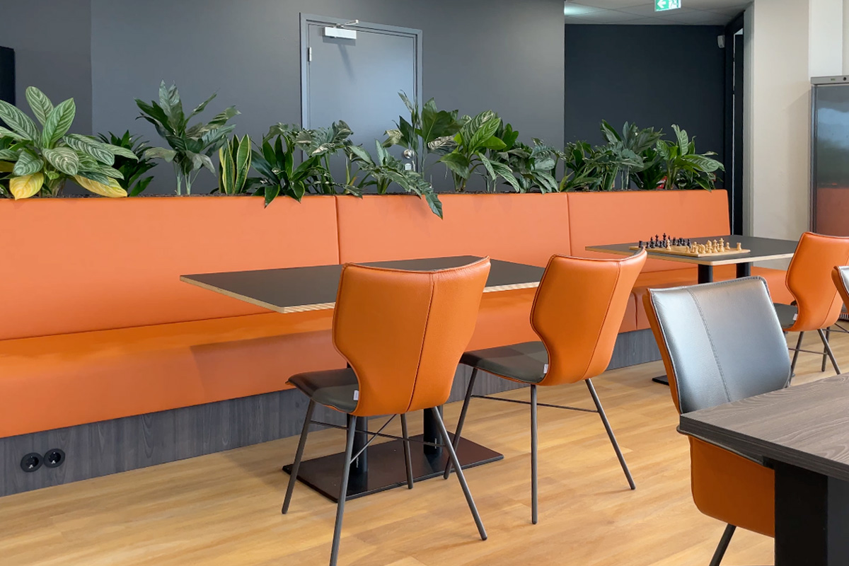 Kantine met maatbank met planten en tafels en stoelen in de oranjekleur van de huisstijl.