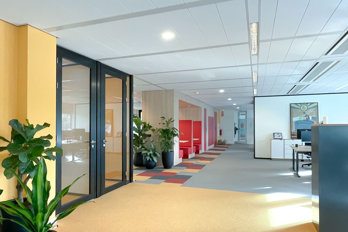 Open kantoorruimte met glaswanden, kleurrijk tapijt, planten en ingebouwde rode zitplekken langs de muur.