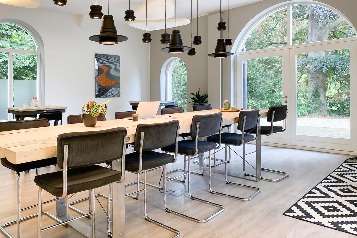 Sfeervolle meetingruimte in het monumentale pand van Vasco met lange houten tafel, zwarte hanglampen en hoge boogramen.