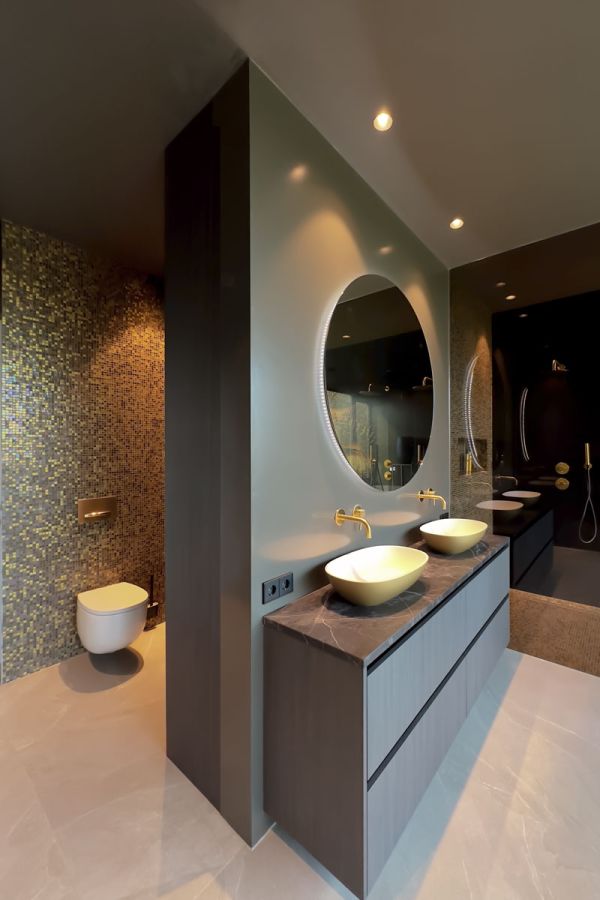 Luxe badkamer met gouden wandtegels, dubbele wastafels op een marmeren blad, en een moderne toiletopstelling. Elegante ronde spiegel met ingebouwde verlichting.