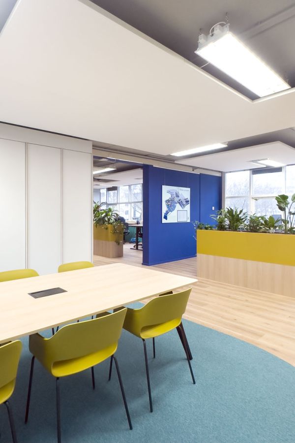 Kantoortuin met vergadertafel, planten, gele stoelen en een blauwe muur.