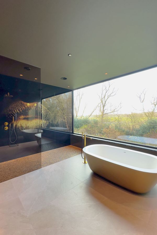 Luxe badkamer met vrijstaand bad, gouden kranen, en een grote glazen wand die uitkijkt op een groene omgeving. Moderne inloopdouche met unieke gouden details.