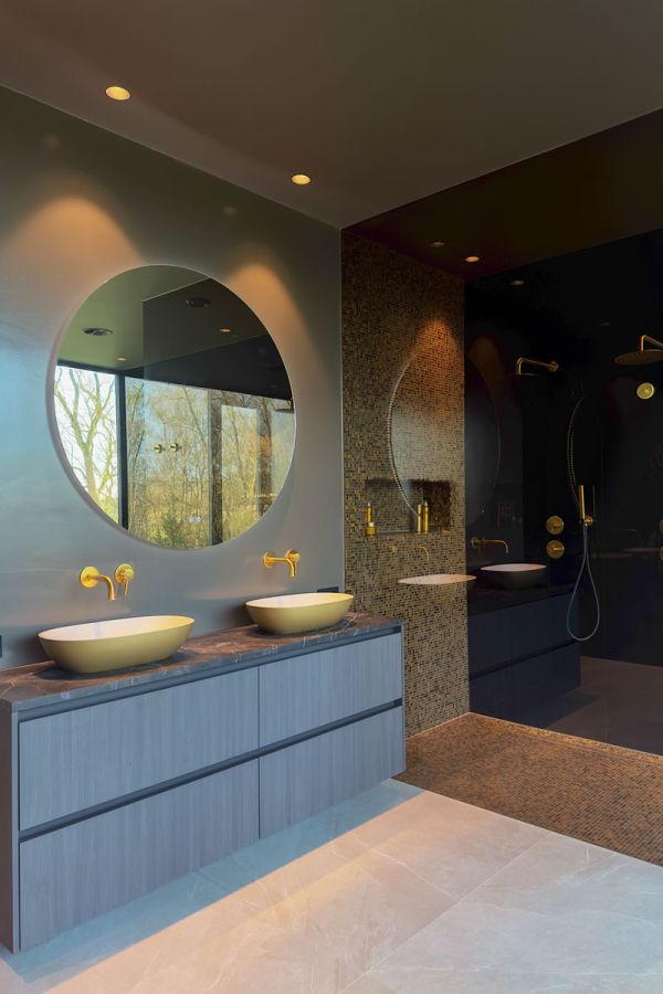 Exclusieve badkamer met gouden wastafels, ronde spiegel met verlichting, en gouden wandkranen. Luxe inloopdouche met mozaïektegels en moderne afwerking.