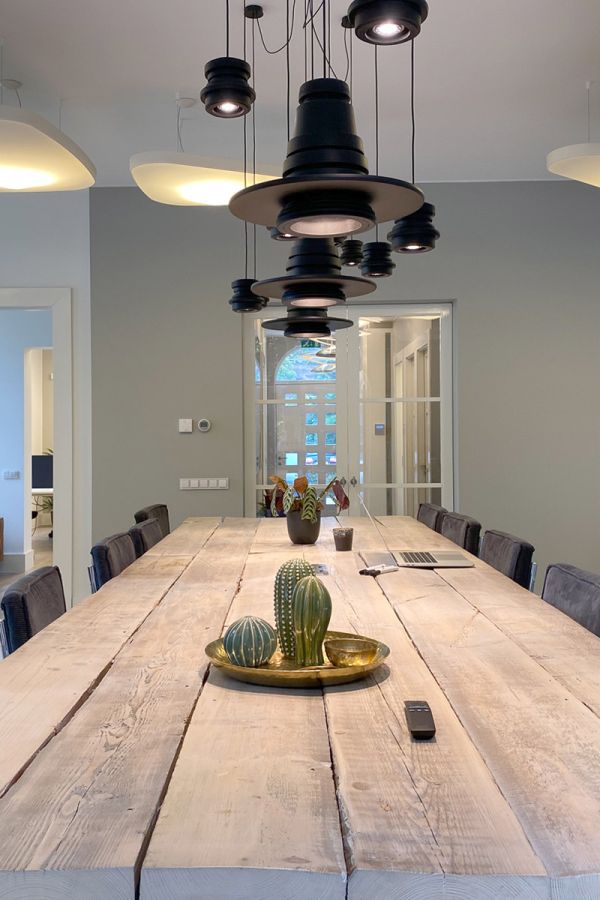 Vergaderruimte met een lange houten tafel, zwarte stoelen en industriële hanglampen. Glazen deuren leiden naar een aangrenzende ruimte.