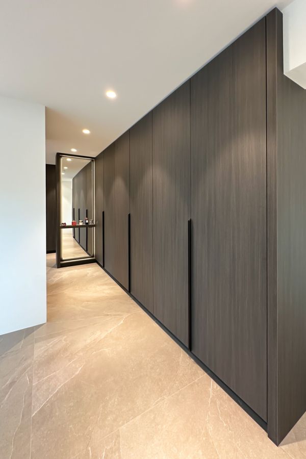 Luxe kledingkast op maat met donkere houten panelen, moderne handgrepen, en een grote spiegel. Voorzien van subtiele ingebouwde verlichting.